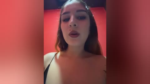 A video of a young woman with light skin, long brown hair, and full lips, wearing a black strapless top, set against a red background.