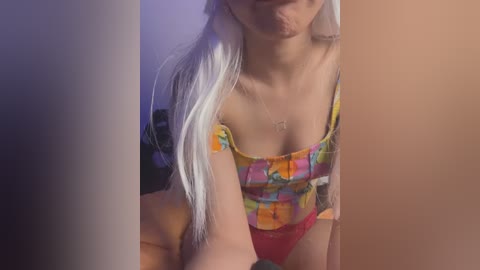 Video of a slender, light-skinned woman with long, platinum blonde hair, wearing a colorful, off-shoulder crop top and red panties. She's in a dimly lit room with purple lighting.