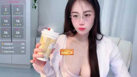 Video of an East Asian woman with long black hair and glasses, wearing a white lab coat, holding a DINGA cup. Background includes a calendar and white curtains.