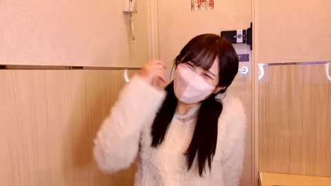 A video of an East Asian woman with long black pigtails, wearing a white fuzzy robe and a white face mask, dancing with her arms raised in a bathroom with light-colored wooden walls and a glass shower door.