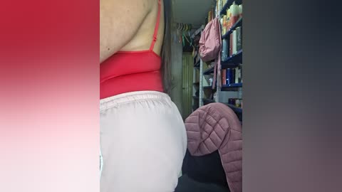 Video of a person with fair skin, wearing a red tank top and white sweatpants, standing in a cluttered room with a pink quilt and a bookshelf visible in the background.