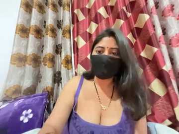 Video of a South Asian woman with medium skin tone, wearing a black face mask, purple tank top, and gold necklace, sitting in front of floral-patterned curtains.