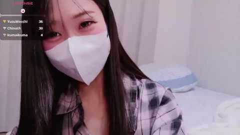 Video of an Asian woman with long black hair, wearing a white face mask and plaid shirt, indoors with white walls and bed.