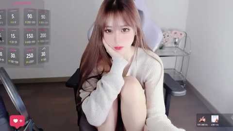 Video of a young East Asian woman with fair skin, long brown hair, and bangs, sitting in a chair in a dimly lit room, wearing a white sweater and a black skirt.