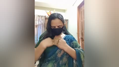A video of a South Asian woman with long dark hair, wearing a blue saree, black mask, and touching her chest, standing indoors in a dimly lit room with wooden furniture and curtains in the background.