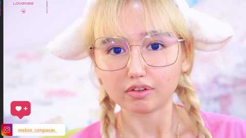 Video of a young woman with fair skin, blue eyes, blonde hair in braids, wearing cat ears and glasses, in a pink top, with a blurred background.