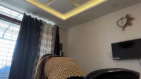 Video of a modern living room with a beige armchair, grey curtains, and a mounted TV on a white wall with a decorative heart-shaped metal piece.