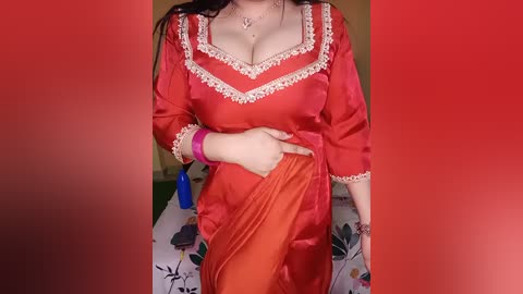Video of a fair-skinned woman with long dark hair, wearing a red, embroidered, traditional Punjabi dress, with a floral tablecloth in the background.