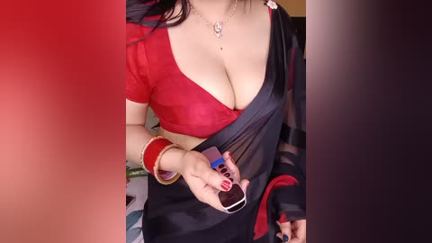 Video of a fair-skinned woman in a red blouse with a plunging neckline, partially exposing ample cleavage. She wears a black sari with a red border, adorned with gold bangles and a silver necklace. Background includes a blurred red and black gradient.