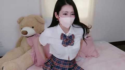 A video of an Asian woman with long black hair, wearing a white blouse, plaid skirt, pink bow, and face mask, seated on a white bed with a large teddy bear.