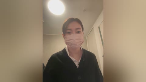Video of a young Asian woman with short dark hair, wearing a black cardigan over a white shirt, and a pink surgical mask. She stands in a dimly lit hallway with beige walls and a ceiling light.