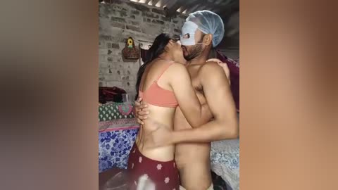 Video of a man and woman in a bedroom, the man shirtless with a blue surgical mask and a woman in a pink bra and maroon pants.
