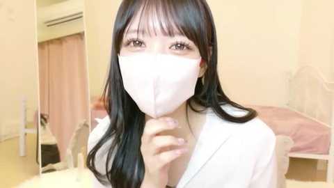 Video of an Asian woman with long black hair, wearing a white face mask and robe, standing in a beige, sparsely furnished bedroom with a bed and pink curtains.