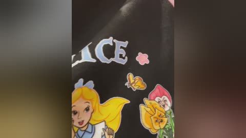 Video of a black surface with a colorful Alice in Wonderland-themed sticker featuring Alice, a rose, and a butterfly, set against a blurred, dark background.