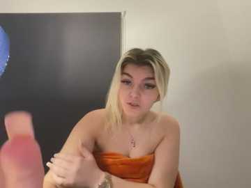 Video of a young, fair-skinned, blonde woman with medium-sized breasts, wearing an orange strapless dress, sitting in front of a black wall, holding hands with a person off-frame.