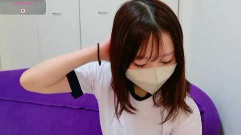 Video of an East Asian woman with straight, shoulder-length dark brown hair, wearing a white T-shirt, beige face mask, and black bracelet, adjusting her mask in a white-walled room with a purple couch.