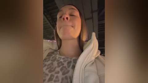 Video of a young woman with light skin and straight, shoulder-length brown hair, smiling blissfully. She wears a leopard-print top and a white jacket. The background is blurred, suggesting an indoor setting.