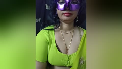 Video of a woman with long black hair, wearing a bright green top, purple mask, and butterfly earrings. She has a fair complexion and medium-sized breasts.