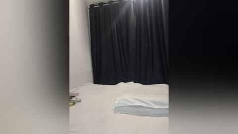 Video of a dimly lit bedroom with white walls and a black curtain covering a window. A neatly made bed with white sheets and a light blue blanket is visible, along with a small, cluttered nightstand with a lamp and a few objects.