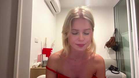 Video of a blonde woman with fair skin and medium-length hair wearing a red off-shoulder top, standing in a modern, minimalist bathroom with white walls, a glass shower, and a wooden dresser holding toiletries.