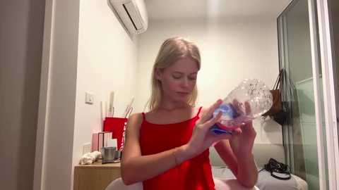 A video of a blonde young woman in a red sleeveless top, filling a plastic bottle with water from a bathroom sink. The room has white walls and a glass shower door.