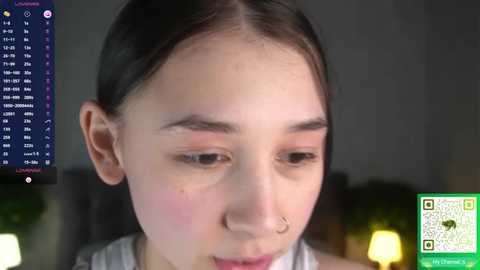 Video of a young Asian girl with fair skin, dark hair, and freckles. She has a nose ring and wears a striped top. Background shows a dimly-lit room with green QR codes on the right.