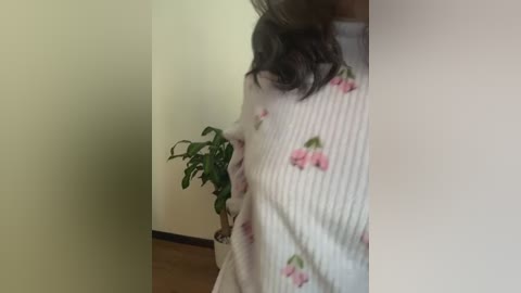 A video of a woman in a white sweater adorned with pink and green floral patterns, holding a potted plant, standing in a beige-walled room with wooden flooring.