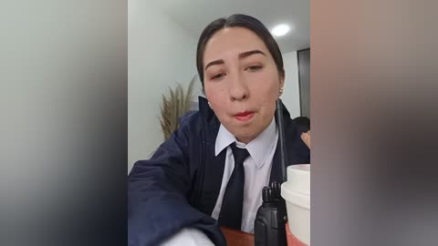 Video of a woman with fair skin, brown hair, and glasses, dressed in a navy blue blazer over a white shirt, holding a coffee cup in a modern indoor setting.