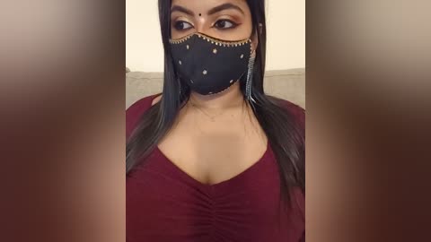 Video of a South Asian woman with medium skin tone, dark hair, and black face mask, wearing a burgundy, low-cut top, sitting against a beige background.