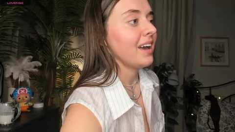 Video of a smiling, light-skinned woman with long brown hair, wearing a white blouse, in a cozy, dimly lit room with potted plants and framed art.