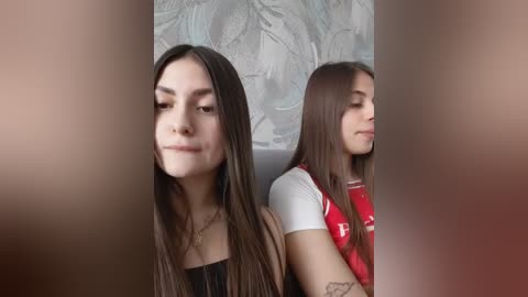 Video of two young women with long brown hair, one wearing a black top and the other a red t-shirt, sitting side by side, against a textured, light-colored wall.