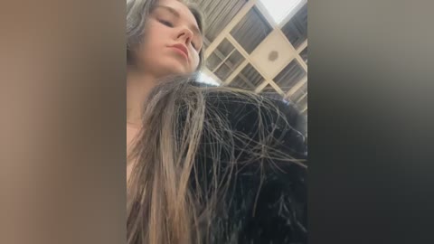 Video of a young woman with long, wet, dark brown hair, wearing a black, shiny jacket, looking down with closed eyes, in a modern, light-filled room with a grid-patterned ceiling.