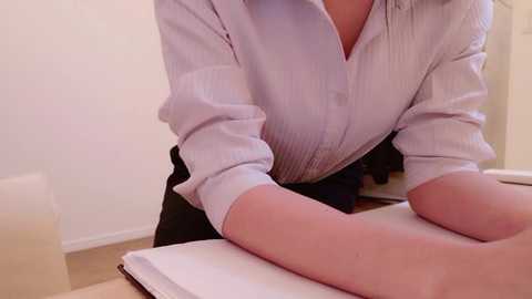 Video of a light-skinned person in a white, striped button-up shirt leaning over a massage table, with a black skirt visible, in a bright, minimalistic room with white walls and a beige floor.