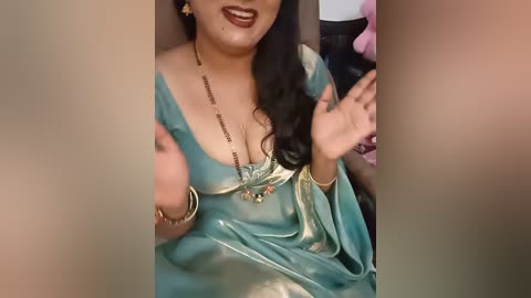 Video of a South Asian woman with medium brown skin, wearing a teal saree, gold jewelry, and makeup, hands raised, indoors.
