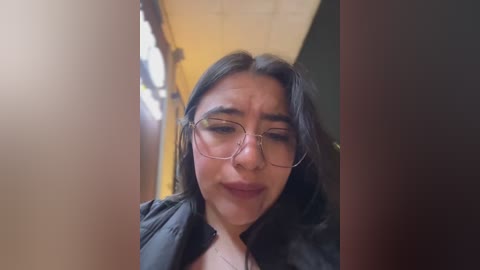 Media: Video of a woman with long dark hair, wearing glasses and a black jacket, looking down with a slight smile, standing in a dimly lit hallway with yellow walls.