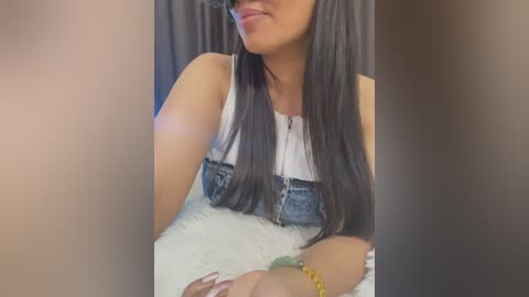 Video of a woman with long, straight black hair, wearing a white tank top with denim straps, sitting on a white fluffy surface.