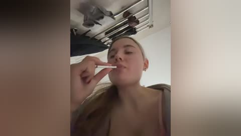 A video of a young woman with fair skin and blonde hair, topless, using a toothbrush to brush her teeth in a dimly lit bathroom with a white ceiling and metallic vent.