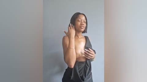 Video of a smiling Black woman with medium brown skin, straightened black hair, wearing a black satin gown that reveals her bare breasts. She stands against a plain, light gray wall, her hands touching her chest.