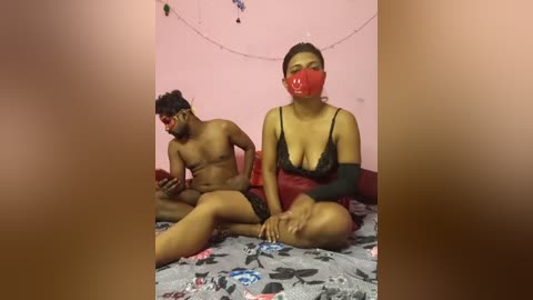 A video captures a nude man with a large penis, sitting beside a clothed woman with red tape over her mouth. Both are on a floral-patterned bed in a pink-walled room with string lights.