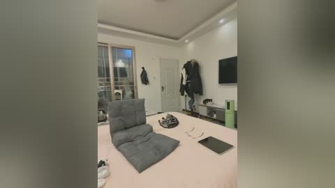 A video of a modern, minimalist bedroom with a grey futon, black coat hanging on a wall, and a flat-screen TV mounted above a green storage unit.