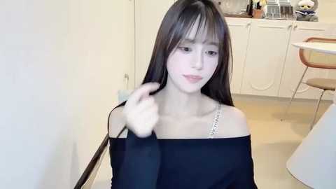Video of an East Asian woman with straight black hair, fair skin, and bangs, wearing a black off-shoulder top, sitting in a modern kitchen with white cabinets and a round table.
