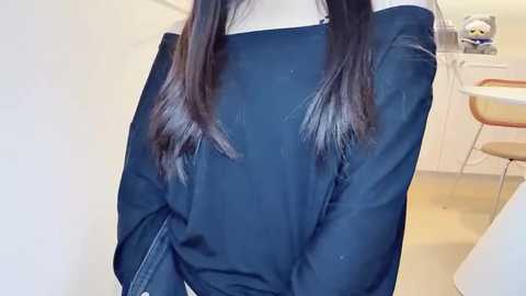 Video of a person with long, straight, dark brown hair, wearing a black, long-sleeved top, standing indoors in a minimalist kitchen with white cabinets, a round table, and wooden chairs.