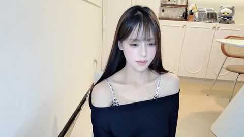 Video of a slender, fair-skinned woman with long, straight black hair and bangs, wearing a black off-shoulder top with a white lace bralette underneath, seated in a modern kitchen with white cabinets, stainless steel appliances, and a round table.