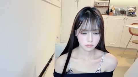 Video of an Asian woman with long black hair and bangs, wearing a black off-shoulder top, sitting in a modern, white kitchen with cabinets and a bar stool.