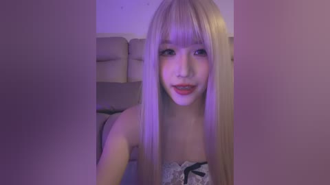 Video of a young, light-skinned woman with long, straight platinum blonde hair and bangs, wearing a white lace bra with black ribbon straps, smiling softly. She is in a dimly lit, beige-colored room with a large, cushioned headboard visible in the background.