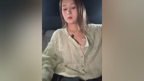 A video captures an Asian woman with straight, shoulder-length brown hair and fair skin, wearing a cream-colored, textured cardigan and a black bra. She appears to be in a dimly lit room, leaning against a dark wall.