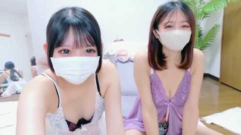 Video of two Asian women with short black hair, wearing white face masks, and revealing lace lingerie, seated on a couch in a modern, brightly-lit room.