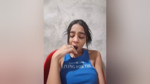 Video of a young Latina woman with medium skin tone, dark hair, wearing a blue sleeveless top that reads \"I'm living for the drama.\" She sits against a textured gray wall, with a red chair in the background.