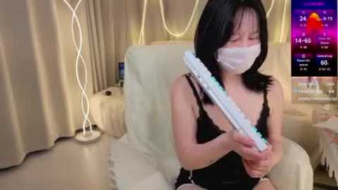 A video of an Asian woman with straight black hair, wearing a black dress, white mask, and holding a blue toy gun, sitting in a beige chair with a white blanket, in a dimly lit room with a TV showing a weather forecast.