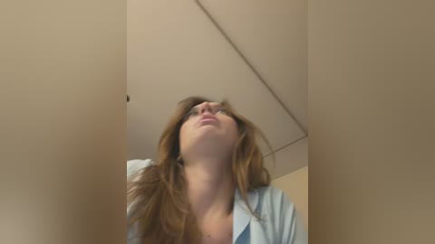 Video of a Caucasian woman with long brown hair, wearing a light blue shirt, standing in a small, beige-walled room with a ceiling fan. She has a serene, slightly tilted head, gazing upward.